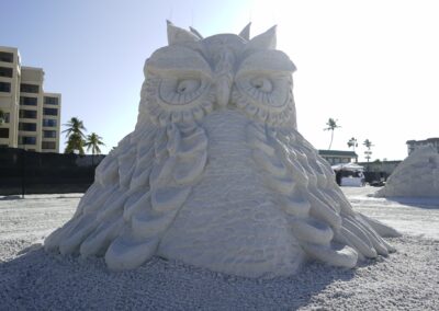 Large sand sculpture of an owl with carved feathers and expressive eyes at a beach festival.