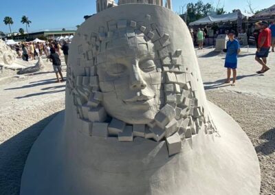 Large sand sculpture of a human face emerging from block-like texture beneath tall cathedral-style spires.