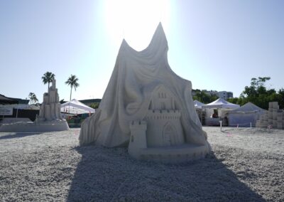 Sandcastle partially covered by flowing draped design with detailed arches and towers at a beach festival.