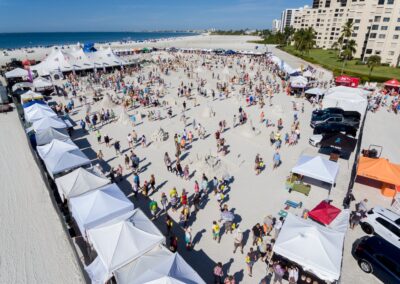 Busy beach festival with rows of white tents, crowds, and multiple sand sculptures near the ocean.