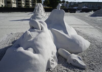 Large sand sculpture of a reclining face beside a serpent-like form at a beach festival.