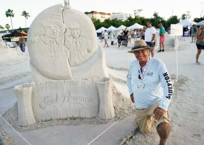 Sand sculpture titled “Soul Reunion” with two figures embracing, displayed beside the sculptor at a beach event.