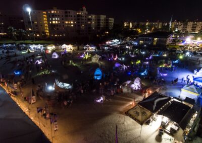 Color‑lit sand sculptures at a nighttime beach festival with crowds and tents in the background.