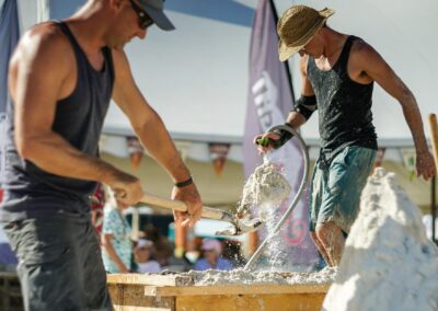 Two sculptors shaping wet sand on a platform using shovels and tools during a beach event.