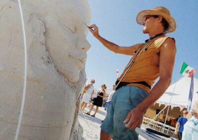 Artist carving a large serene face in sand, adding details under a sunny sky at a festival.