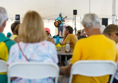 Musician performing with an acoustic guitar under a tent while attendees watch from tables.