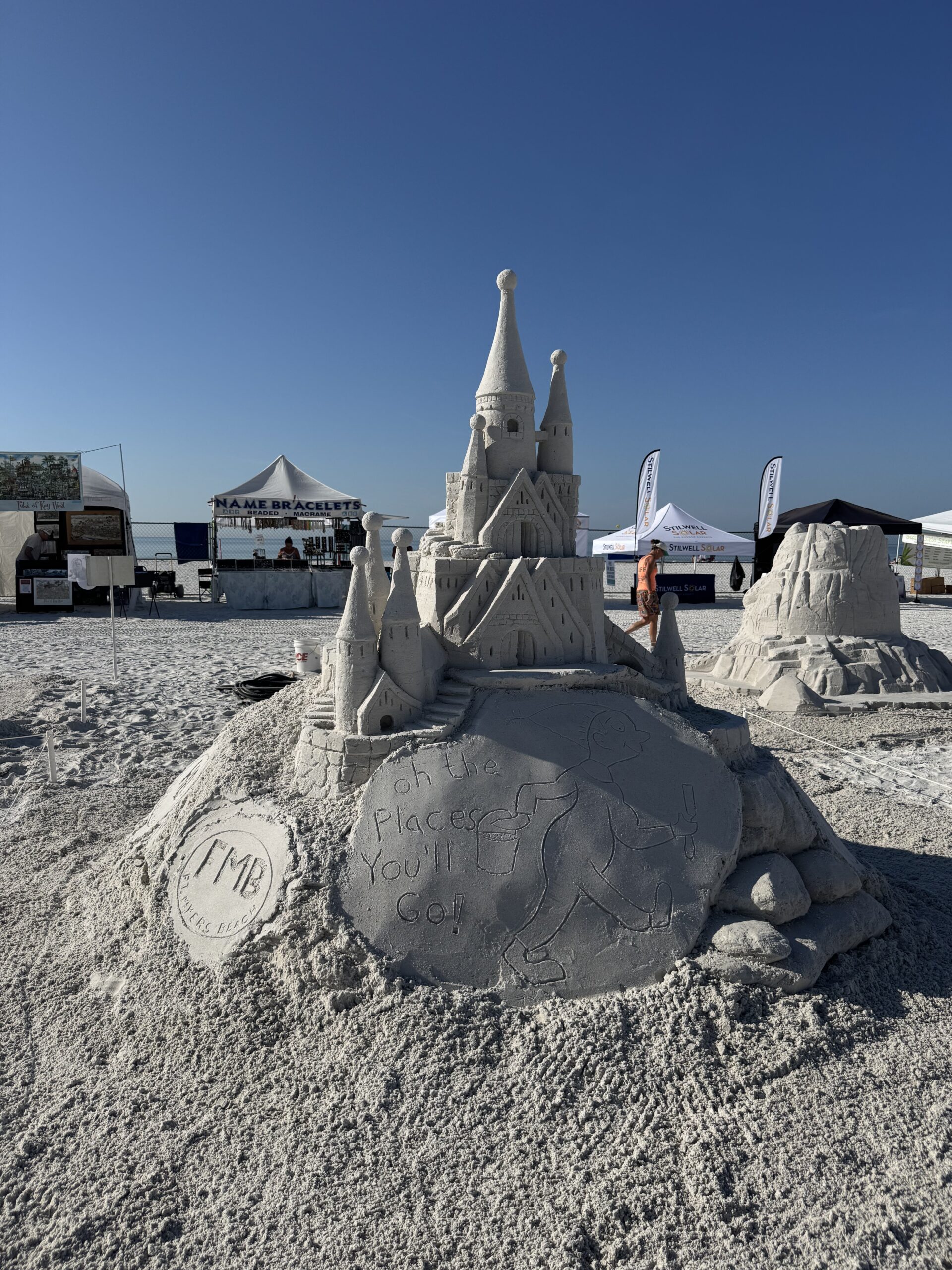 darrell o'conner Sandcastle with “Oh the Places You'll Go!” plaque at sunny beach fest, tents in background.