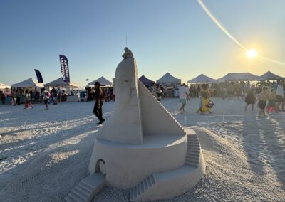 Tower-like sand sculpture with face and hand on top, surrounded by crowd and tents at sunset.