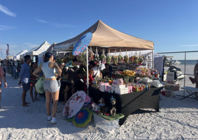 Beach market tent with plush toys, flowers, and “Anna’s Handcrafted Creations” sign, jet ski in background.