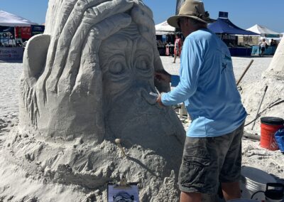 Artist carving a detailed sand sculpture of a face with hands on the forehead at a beach event.