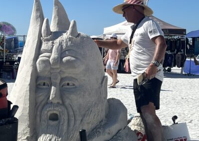 Sculptor shaping a large sand sculpture of a bearded head with tall horn‑like structures.