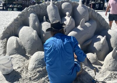 Artist carving a circular sand sculpture of multiple dolphins emerging from the sand at a beach festival.