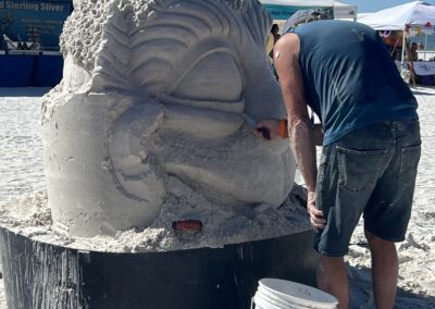 Person carving a detailed sand sculpture shaped like a stylized fish head at a beach event.