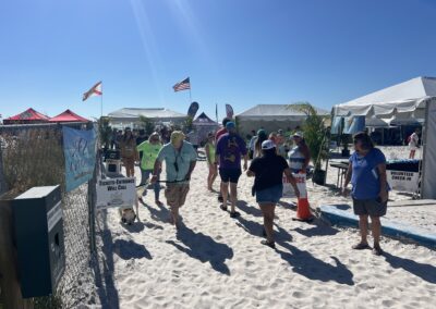 People walking on white sand near event tents with entrance signs, flags, and sponsor banners.
