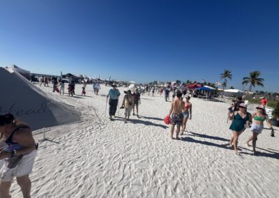 Crowds walking along a sunny beach lined with tents and palm trees during a lively festival.