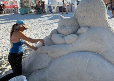 Artist carving a sand sculpture of a child hugging a cat at a beachside art event.