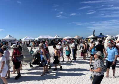 “Busy beach festival with people walking past tents and sand sculptures, including a carved rose.”