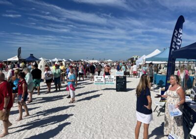 “Crowded sandy walkway lined with tents and a sign for restrooms and live music under a clear sky.”