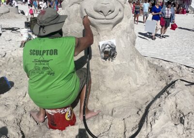 “Sculptor in a green shirt carving a smiling lion face into a large sand sculpture using a reference photo.”