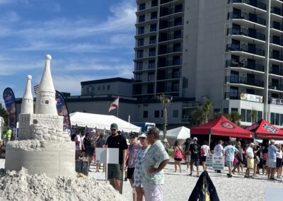 “Detailed multi‑tower sandcastle on the beach with spectators and tents in front of a tall building.”