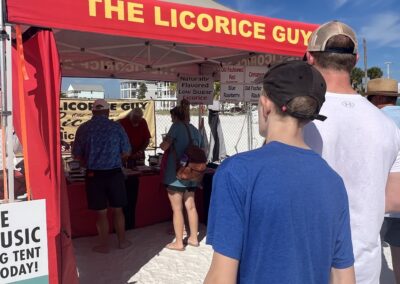 Red-and-tan “The Licorice Guy” tent with people lined up on sandy ground at an outdoor event.