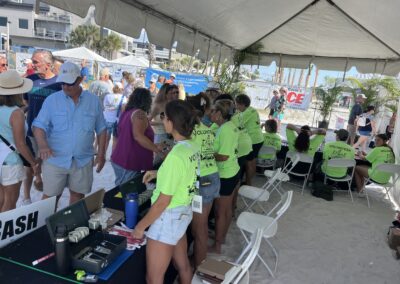 Volunteers in neon shirts assist attendees under a large white beach tent with sponsor banners behind them.