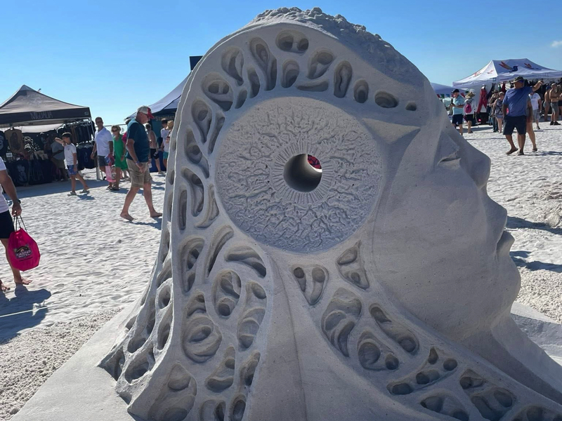Sand sculpture of a stylized human face in profile with flowing carved patterns and a circular hollow feature.