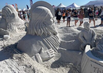 Large sand sculpture of a bearded figure beside a small boat at a beach festival with tents and crowds.