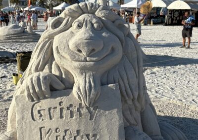 Cartoon-style sand lion holding a “Gritty Kitty” sign with a football in front at a beach festival.
