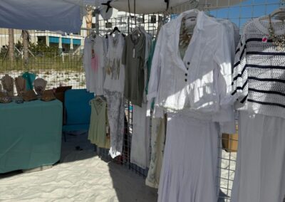 Beachside vendor tent displaying white clothing and jewelry on a sandy lot with buildings in the background.