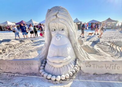 Humorous sand sculpture of a wide-eyed character with signs pointing to “The Wife” and “Husband Around Back.”