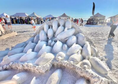 Large sand sculpture of multiple dolphins rising from a mound on a sunny beach with event tents and visitors in the background.