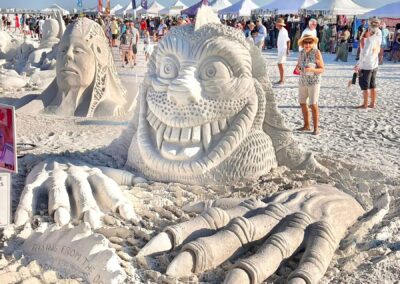 Sand sculpture of a grinning sea creature with large hands and the carved text “RISING FROM THE DEEP,” with another face sculpture and event tents behind it.