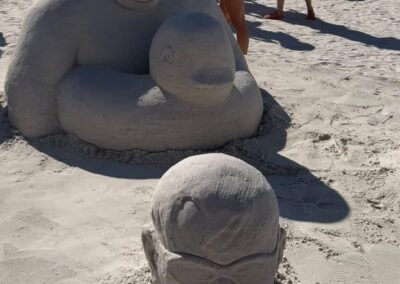 Sand sculpture of a large expressive figure behind a smaller head wearing sunglasses, with a sand duck float and people walking among event tents.