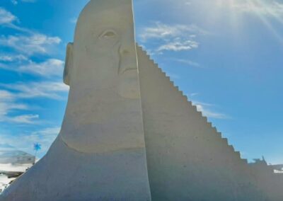 Tall sand sculpture of a serene human face combined with stepped geometric shapes, set against a bright sky with scattered clouds.