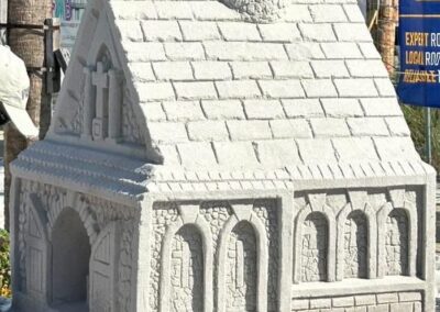 Detailed sand sculpture of a small chapel with a pitched roof, arched windows, and a cross carved into the front gable.