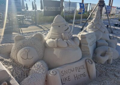 Sand sculpture of festive characters and signs on a beach with sponsor banners behind it.