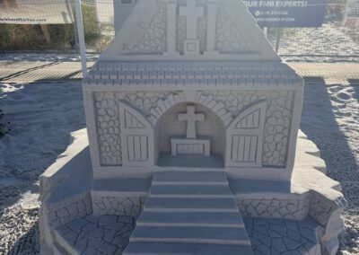 Detailed sand sculpture of a small chapel with carved crosses and sponsor banners in the background.