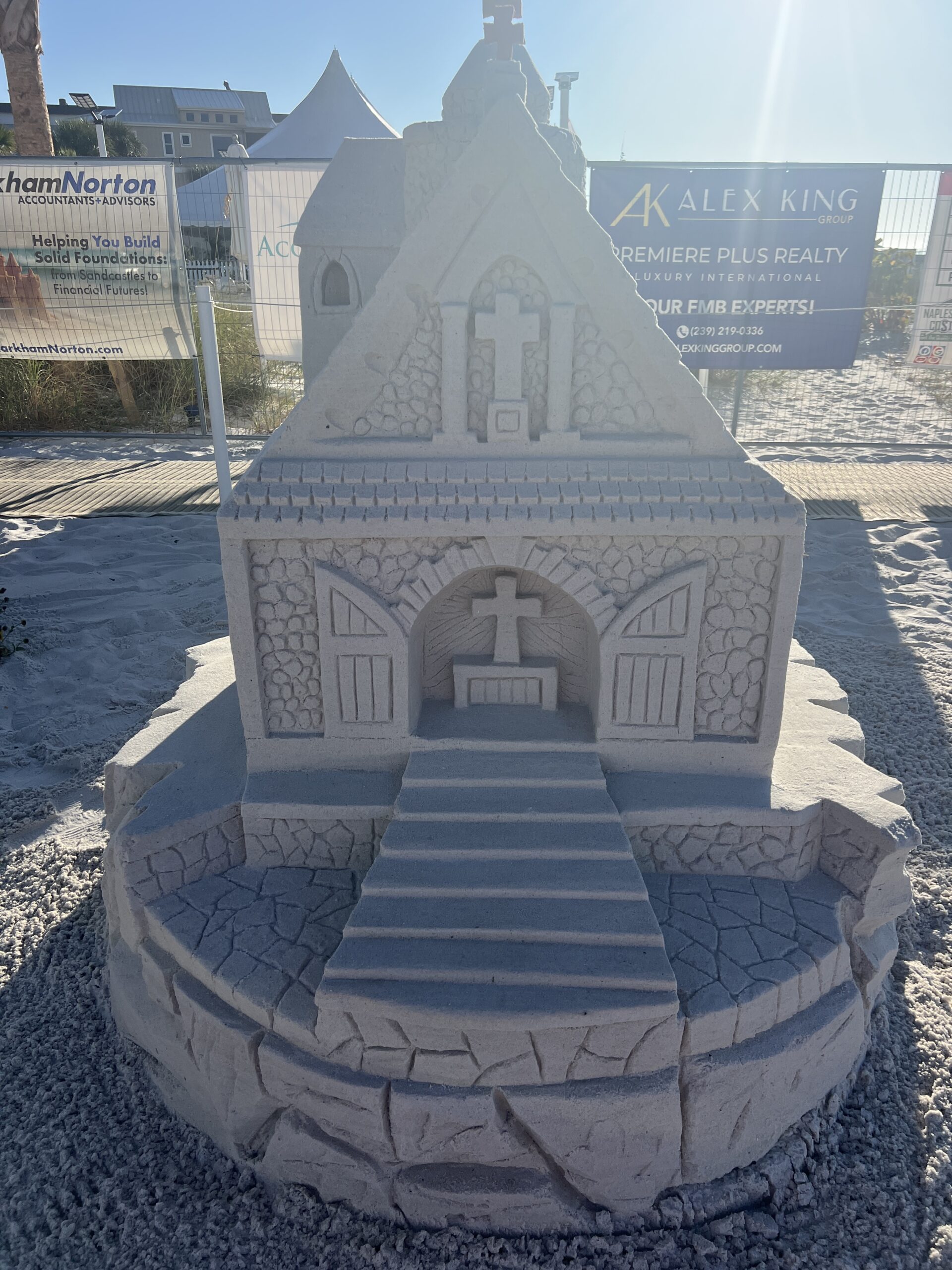 IMG_0903 Detailed sand sculpture of a small chapel with carved crosses and sponsor banners in the background.