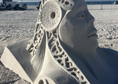 Large abstract sand sculpture of a stylized human head on a beach under a clear sky.