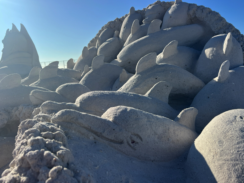 Detailed sand sculpture of multiple dolphins emerging together from a mound, set against a clear sky.