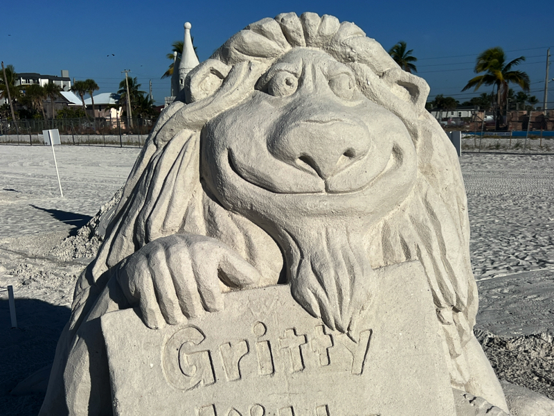 Sand sculpture of a smiling creature with long hair holding a plaque labeled “Gritty,” set on a beach.