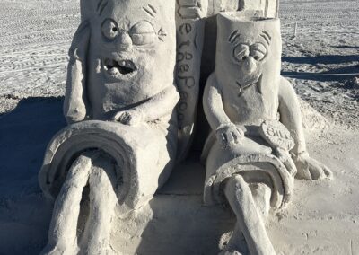 Sand sculpture of two expressive rectangular characters sitting in front of a tall structure carved with “We the People.”