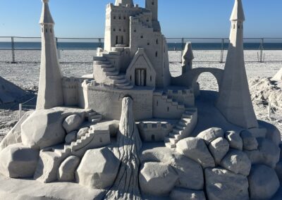 Large sandcastle with tall pointed towers, arches, and a rocky base, set against the ocean and blue sky.
