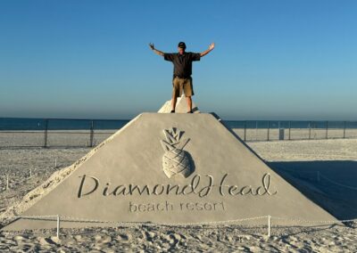 Person standing on a large sand sculpture shaped like a pyramid with “DiamondHead Beach Resort” carved on it.