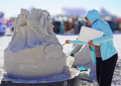 Person in a blue jacket carving an intricate sand sculpture with flowing shapes, using small tools at a beach event.