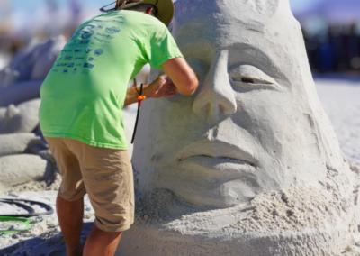 Artist in a green shirt and hat shaping the nose of a large sand face sculpture at a beach competition.