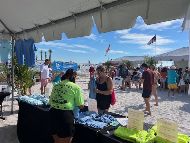 IMG_0735 Volunteers in neon shirts assist attendees under a large white beach tent with sponsor banners behind them.