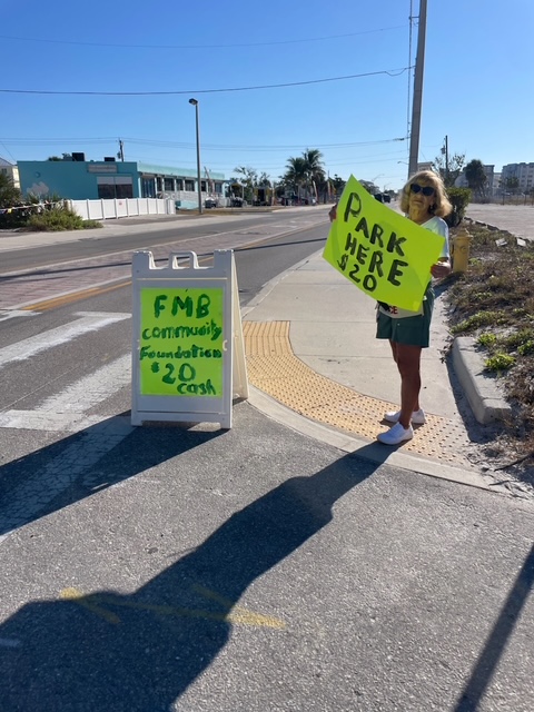 parking-lot Woman holding neon "PARK HERE $20" sign beside matching FMB Community Foundation donation board on sunny street corner.